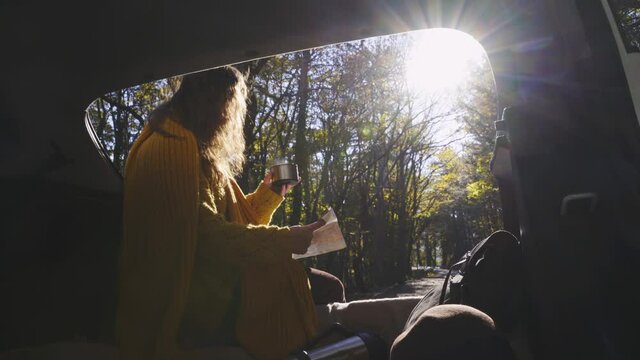 Curly haired woman with yellow scarf drinks tea and reads paper map sitting in open trunk of hatchback car on road in autumn forest backside view