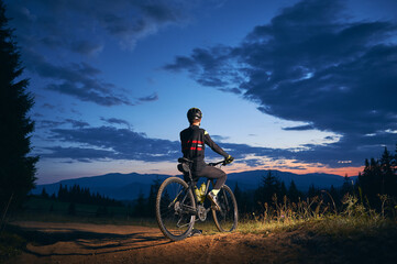 Back view of man sitting on bicycle under blue night sky with clouds. Male bicyclist in safety helmet riding bicycle on the trail in the evening. Concept of sport, biking and active leisure.