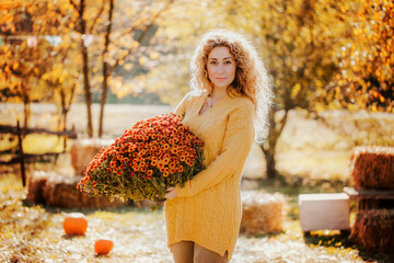 Beautiful curly girl holds an armful of orange autumn flowers against the background of the autumn fair