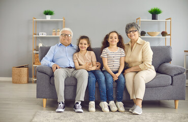 Family group portrait of happy grandparents and little grandchildren at home. Senior man and woman together with cute little children sitting on the sofa in the living-room and looking at the camera