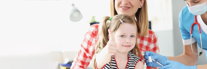 Naklejka premium Little girl holds her thumbs up and doctor vaccinates her shoulder in medina office