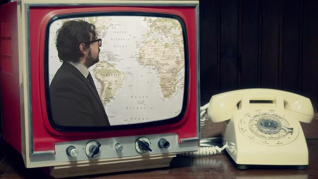 A transmission in an old television set near a rotary phone: a man in an elegant suit pointing at different parts of a world map (weather report).
