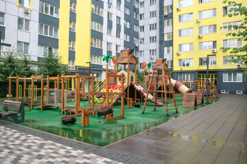 A courtyard of high-rise buildings with a modern and large playground made of wood and plastic on a rainy summer day without people. Empty outdoor playground. A place for children's games and sports.