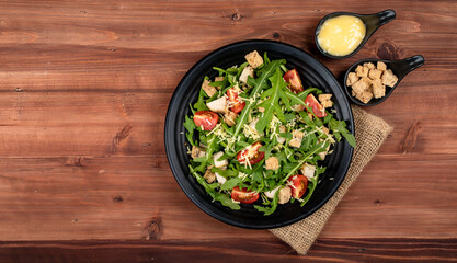 Rocket salad with tomato, crouton, herb roasted chicken, parmesan cheese in black dish on sackcloth together with two tiny sauce cups full of mayonnaise and extra crouton  on the dark wooded table