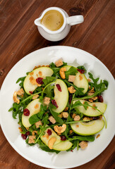 Fresh green apple slice salad with rocket leaf, oat granular, dried cranberry, and cashew nut placed in white dish on the dark wooded table. olive oil with vinegar salad dressing as a side dish