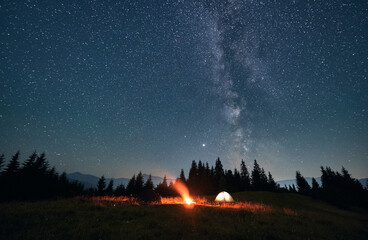 Wide angle view on beautiful landscape in the mountains. Tourist tents and campfire under beautiful starry sky. Mountains range behind spruces. Concept of night camping and astrophotography