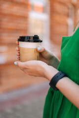 Vertical close-up cropped shot of unrecognizable young woman holding takeaway coffee cup outdoors in city street in summer sunny day on blurred background, selective focus, side view.