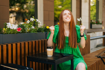 Emotional happy young woman keeping hands in fists, screaming celebrating win, sitting at table with coffee cup and phone in outdoor cafe terrace in summer day, blurred background, selective focus.