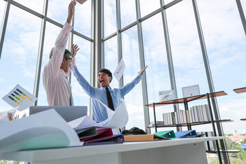 Two Asian professional businessmen celebrating their successful project by throwing document papers in the air with funny happy smiles in the daytime. Concept of achievement of a new start-up company