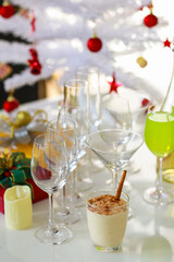 Tasty delicious milk cream eggnog drink with gingerbread in glass on table in front empty tall wine glasses present gift boxes white chocolate candles green apple punch and decorated Christmas tree