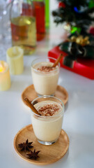Top view shot of two sweet creamy alcoholic eggnog drinks with gingerbread on wooden coaster with anise on white clothing table full of empty tall wine glasses liquor bottles and present gift boxes