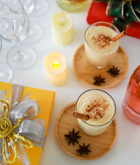 Top view shot of two sweet creamy alcoholic eggnog drinks with gingerbread on wooden coaster with anise on white clothing table full of empty tall wine glasses liquor bottles and present gift boxes