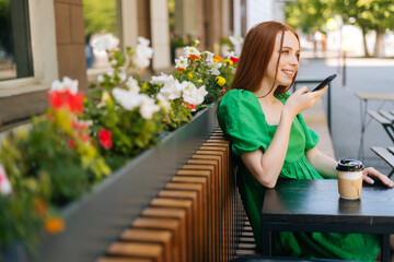 Side view of smiling redhead young woman recording audio voice message on cellphone sitting at table with coffee cup in outdoor cafe terrace in sunny summer day.