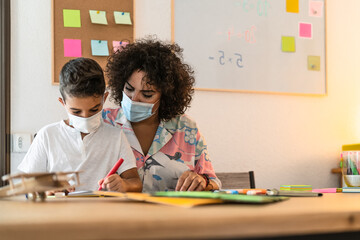 Female teacher with child drawing in preschool classroom while wearing face protective mask during corona virus pandemic - Healthcare and education concept - obrazy, fototapety, plakaty