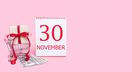 A gift box in a shopping trolley, dollars and a calendar with the date of 30 november on a pink background.