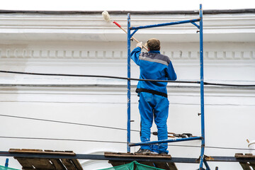 The artist paints the facade of the building with white paint using a roller brush and scaffolding. Beyond recognition. Back view