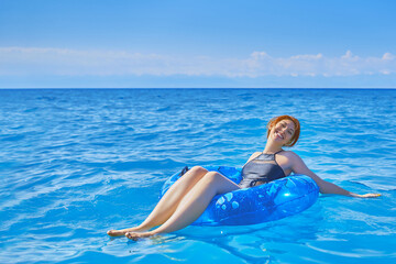 Young red-haired woman floats on ocean in swimming circle. Black fashionable swimsuit. Summer holidays at sea. Beautiful young woman relax on inflatable ring in sea water.