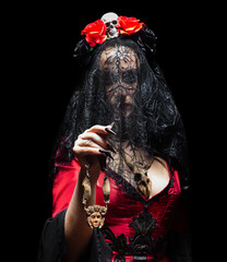 Mysterious woman (sorceress or witch) in red gothic dress, black veil and crown with skull and roses, with two wooden amulets on black background. Halloween concept.