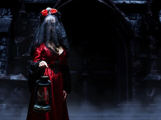 Mysterious witch or sorceress in red gothic dress, black veil and crown with skull and roses holding a lantern standing in front of the church in dark dungeon. Halloween concept.