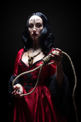 Beautiful brunette sorceress or witch with runic makeup in red gothic dress and wooden animal skull amulet holding a noose on black background. Halloween concept.