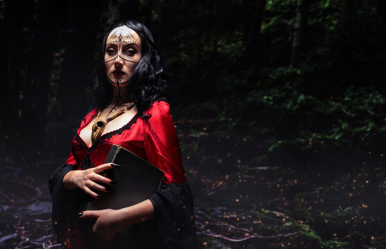 Mysterious sorceress or witch with runic makeup in red dress with wooden animal skull amulet holding a magic book in night fantasy forest. Halloween concept.