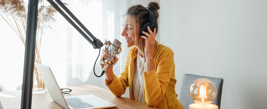 Cheerful woman podcaster recording her voice into microphone. Female radio host streaming podcast using microphone and laptop at his home studio