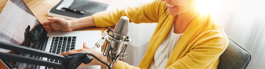 Cheerful woman podcaster recording her voice into microphone. Female radio host streaming podcast using microphone and laptop at his home studio. Wide image - obrazy, fototapety, plakaty