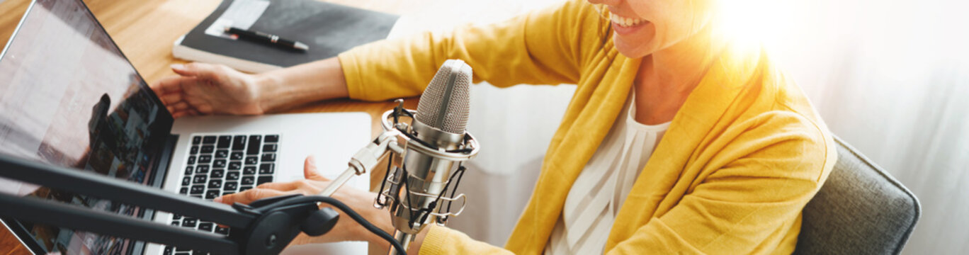 Cheerful woman podcaster recording her voice into microphone. Female radio host streaming podcast using microphone and laptop at his home studio. Wide image