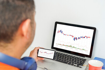 Hispanic man checks on his computer and cell phone the movement of his stocks on the stock market after listening to music