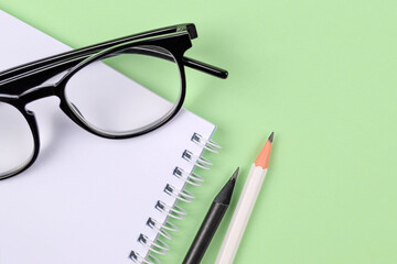 A spring-loaded notebook,white and black pencils,stylish glasses on a delicate green background.Things for businessmen, schoolchildren,students,teachers, artists, education, top view.Copyspace