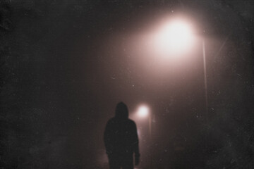 A hooded man standing underneath a street light, back to camera. On a foggy winters night. With a blurred, textured edit.