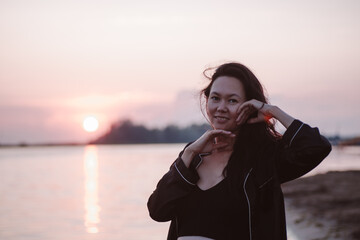 Portrait of woman at sunset. Close-up photo of young smiling Asian woman with long hair on beach and holding her hands her face. 