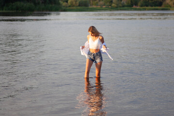 Photo of a woman in white clothes on the background of a river. A beautiful young blonde looking at the water in profile in a white top and shirt and denim shorts stands in the water.