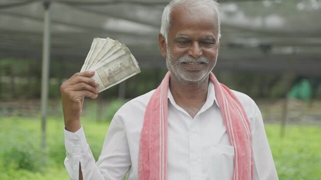 happy smiling Indian farmer holding bunch of Currey notes using and using it as fan at greenhouse or polyhouse - concept of successful profitable agribusiness.