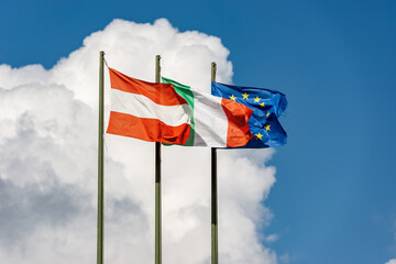 Austrian, Italian and European Union flags with flagpole, blowing togetherness in the wind on a clear blue sky with clouds and copy space.