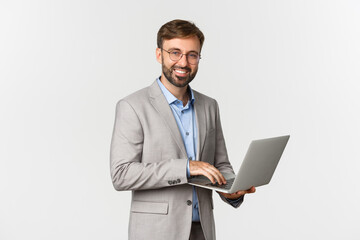 Fototapeta na wymiar Portrait of successful and confident businessman in grey suit and glasses, working with laptop and smiling happy at camera, standing over white background