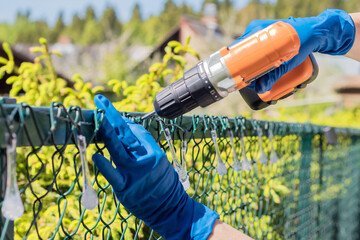 Working with an electric drill with a screwdriver on the mesh of a metal fence of a garden plot. 