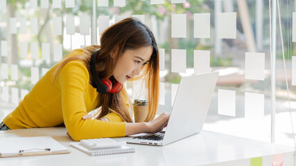 A smiling Asian teen female student in a yellow shirt studying on a laptop doing online research for a report. Take note of the essay homework. e-learning ideas online education at home