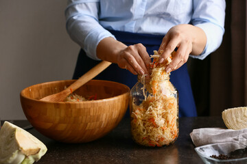 Woman putting tasty sauerkraut into glass jar on table in kitchen, closeup