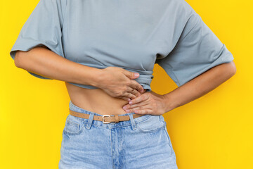 Cropped shot of a young woman in a grey t-shirt and denim shorts holding her side with her hands isolated on color yellow background. Sore side under the rib after training. Stomach pain