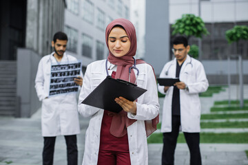 Pretty doctor in hijab writing notes on clipboard, standing ouside modern clinic. Two male Arab hindu doctors standing on the background, using digital tablet and examining patient's xray