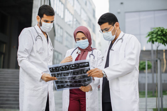 Work in coronavirus pandemic conditions. Three young focused multiracial doctors in masks, standing outdoors in front of modern clinic and discussing patient's tomography scan
