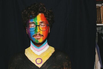 Portrait of young man face painted as lgbtq progress pride flag with direct lighting on face with dark black copy space empty background with flash *3