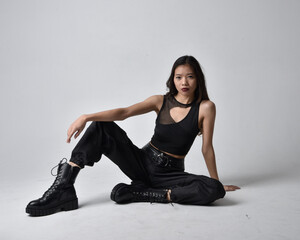 Full length portrait of pretty young asian girl wearing black tank top, utilitarian  pants and leather boots. Sitting poseisolated against a  studio background.