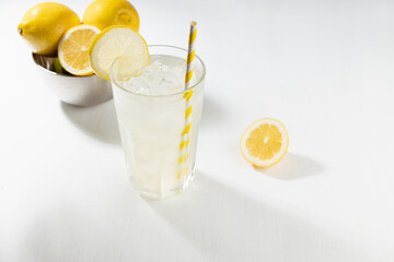 Fresh summer homemade lemonade in misted glass with ice, straw  with ingredient in silver bowl, yellow lemons in sunlight on white wood table.