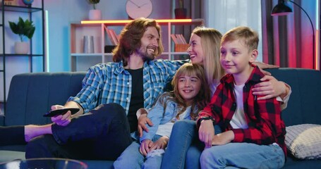 Family idyll of attractive happy smiling loving parents with their two kids sitting together on the couch in the evening and having fun while revisioning show on TV