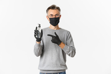 Fototapeta na wymiar Concept of coronavirus, lifestyle and quarantine. Attractive middle-aged man in black medical mask and gloves, recommending use hand sanitizer, pointing at antiseptic, standing over white background