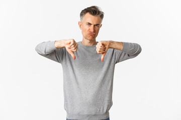 Skeptical and disappointed middle-aged man, grimacing unamused and showing thumbs-down, dislike something bad, standing over white background