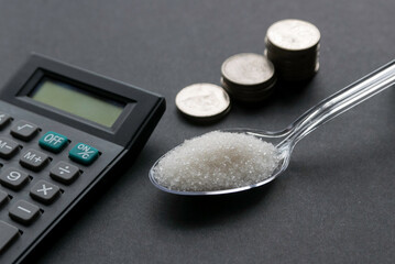 A white sugar in a transparent spoon together with a stack of coins and calculator on a black background.