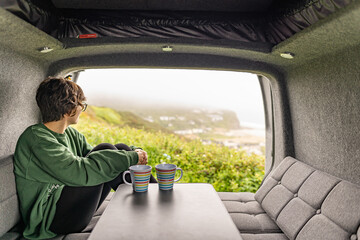 Photo of an attractive young female drinking coffee and sitting on a sofa inside her campervan while enjoying the views on a foggy day